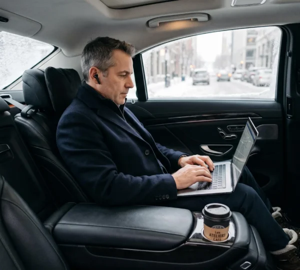 An executive types on a laptop on the rear center armrest of a sedan during a winter ride, with a wool coat folded on the adjacent seat.